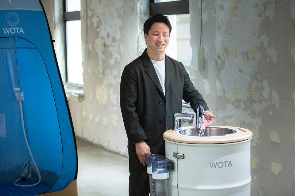 WOTA's CEO MAEDA Yosuke receiving clear water running from a white and round WOSH handwashing stands in a room with unfinished walls.