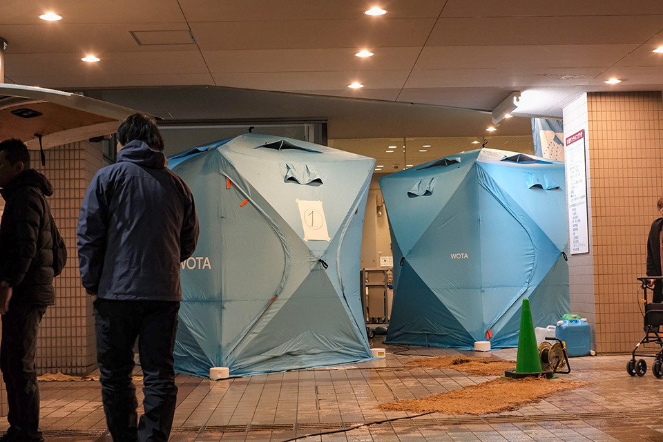 Two people standing in front of two blue tents set up on a tiled floor outside of an facility, with some equipments around.