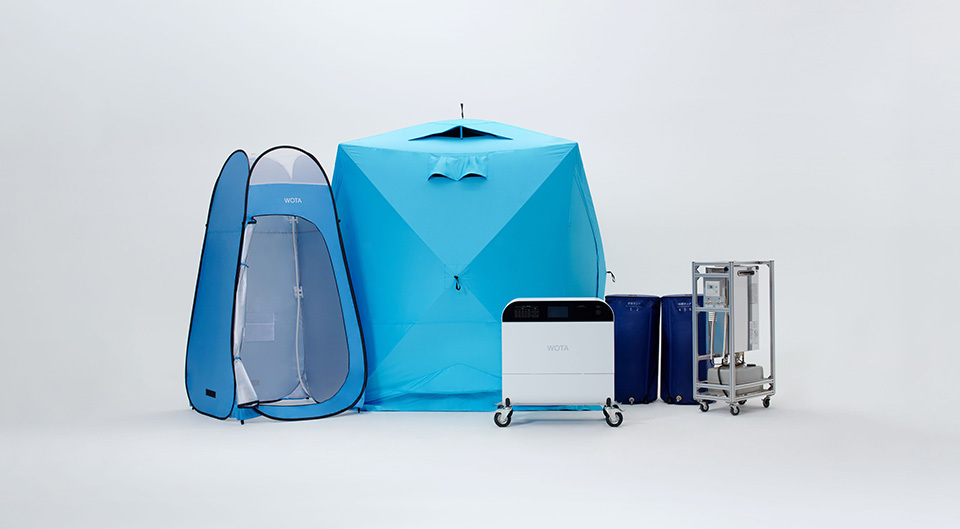 WOTA BOX (wheeled white object in front center) and a collection of equipment on the background, including a blue shower tent, clothes-changing tent, water tank, and water heater.