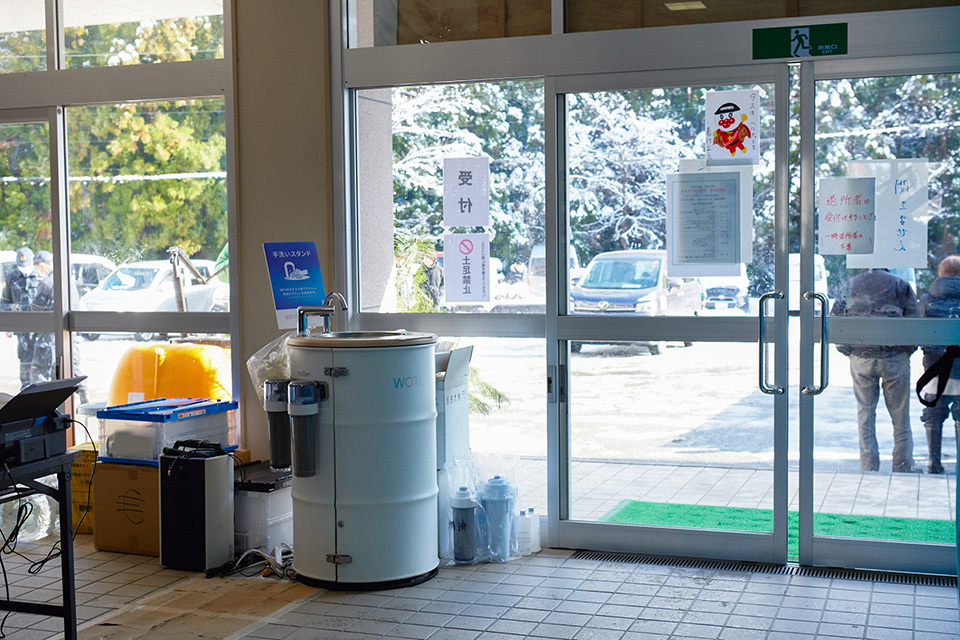 A white cylindrical handwashing stand WOSH is placed near two sets of sliding glass doors at an entrance of an evacuation center.