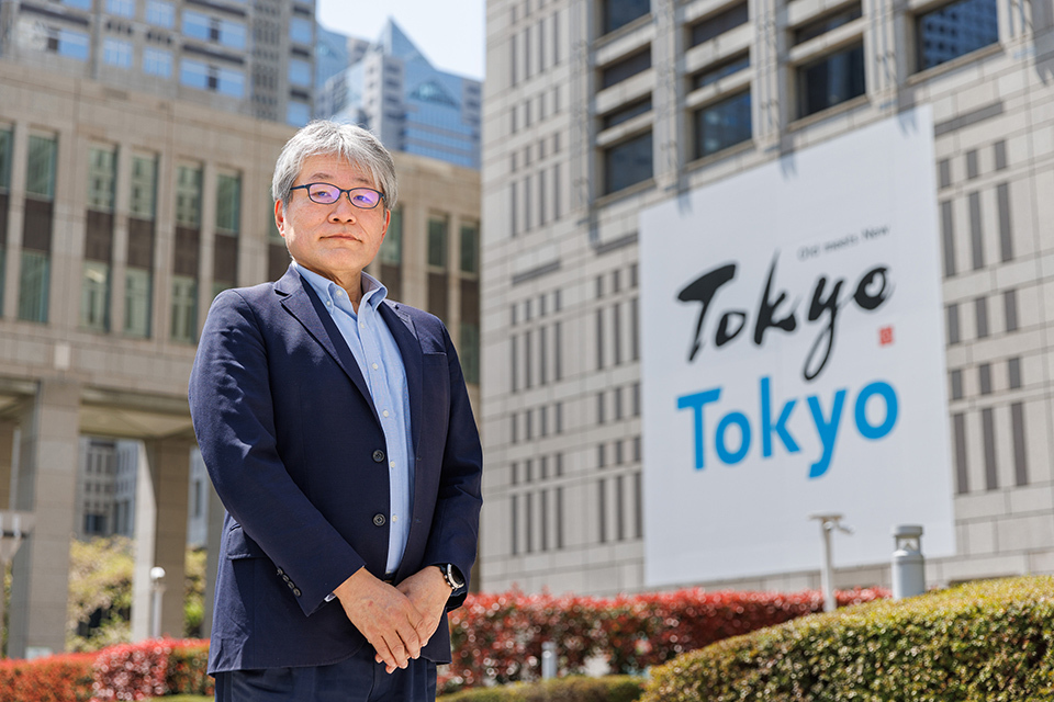 Okamoto Seiji of the Tokyo Metropolitan Bureau of Transportation in a suit poses in front of a white building with a huge rectangular Tokyo banner displayed.