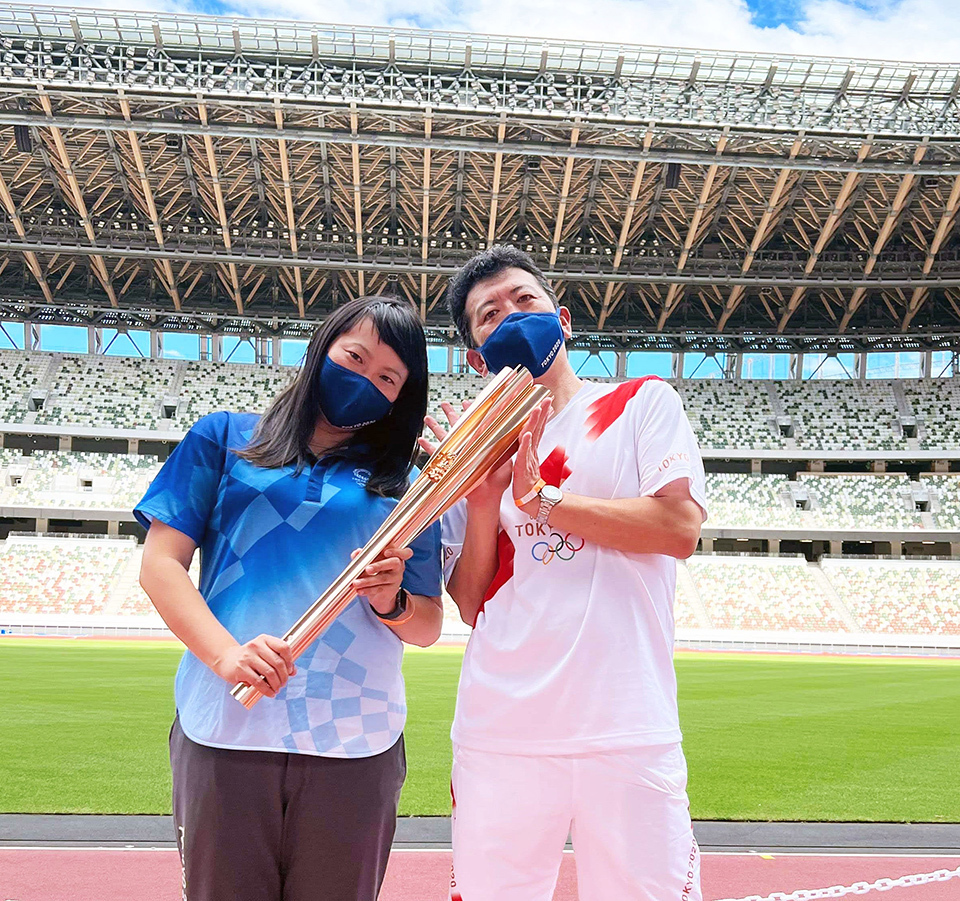 Two individuals holding a large torch together on a track field inside a stadium.