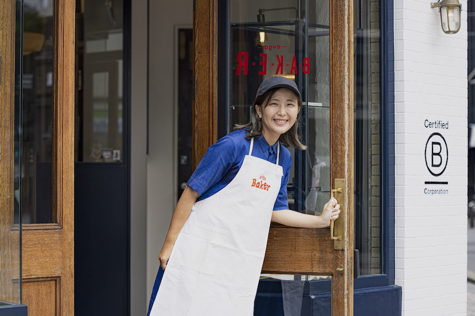 A women in blue apron opens the entrance door of a bakery.