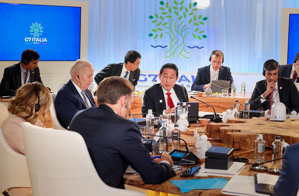 A large wooden circular table with multiple world leaders and officials seated around it