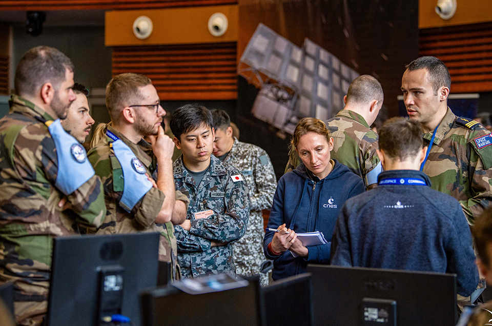 Participants of AsterX, a multilateral tabletop exercise, in military and civilian attire collaborating around computer workstations.