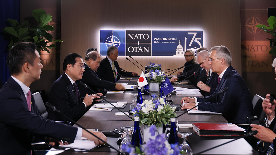 A group of individuals seated around a conference table at a NATO meeting, with the NATO logo visible in the background.