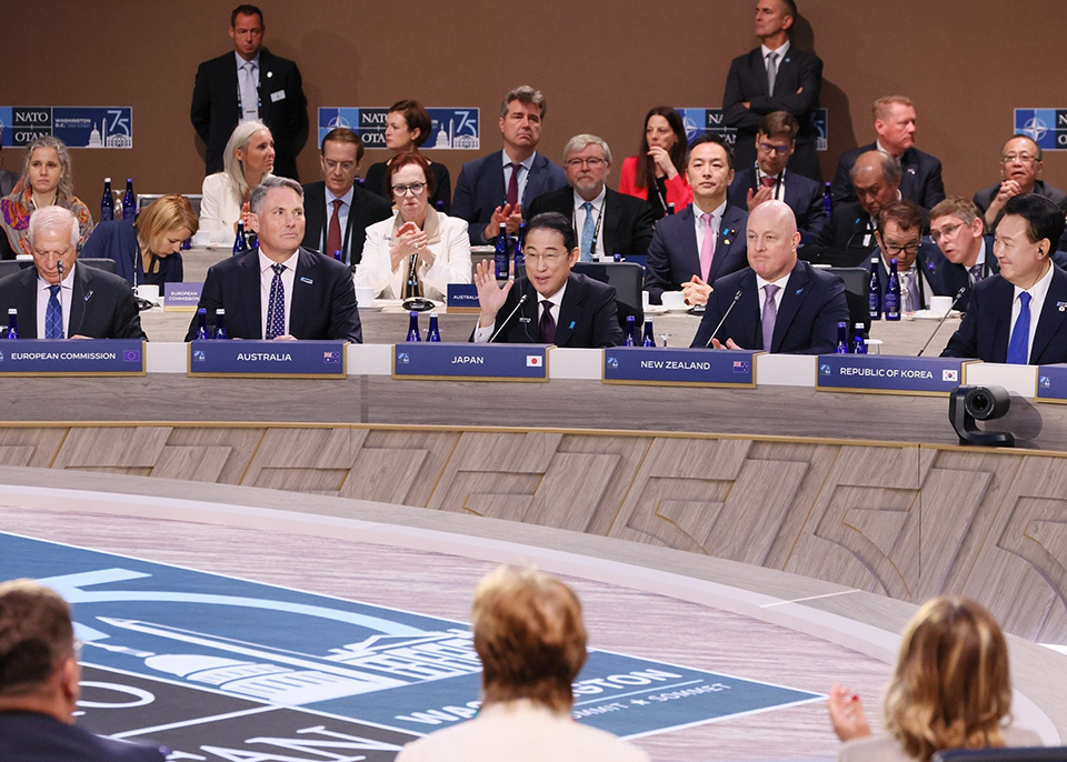 Meeting room at the NATO Summit with participants seated in circular formation around central emblem.