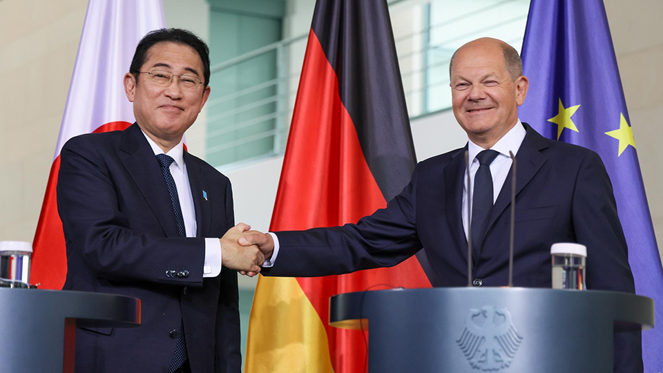 Two individuals engaging in a handshake at a formal event with national flags and an emblematic podium in the background.