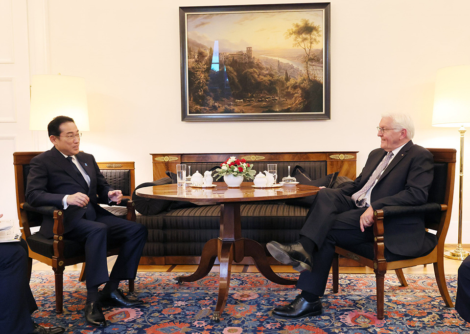 Two individuals sitting opposite each other on sofas in a formal setting, with a coffee table between them.