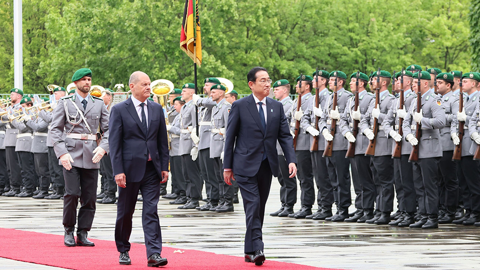 Two men walking on a red carpet, flanked by rows of uniformed personnel standing at attention during an outdoor formal ceremony.