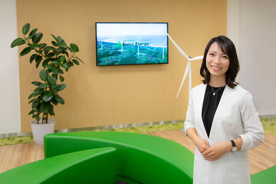 Person in white blazer standing in front of a green sofa with a mounted TV and a potted plant in an office setting.