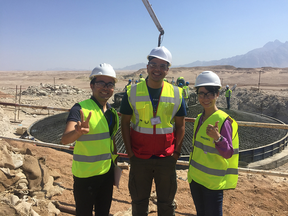 Three people in safety gear give the thumbs up at a construction site where machinery is operating in a dry environment.