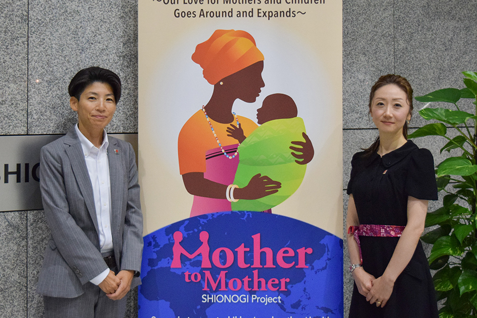 Two women standing beside the "Mother to Mother SHIONOGI Project" stand board.