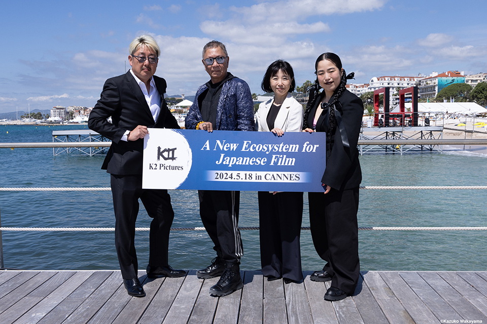 Four individuals standing on a wooden deck by the water in Cannes, holding a sign that reads 'A New Ecosystem for Japanese Film, 2024.5.18 in Cannes.