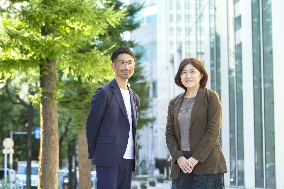 Japanese business professionals Kajiwara and Kanedome standing outdoors, urban background with trees and office buildings