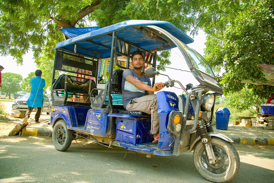 Terra Motors' electric three-wheel auto-rickshaw with driver in India, blue canopy, on sunny street with green trees.