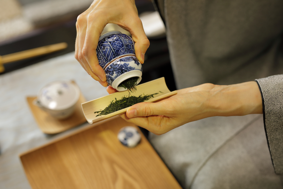 Senchado green tea preparation: Hands pouring loose tea leaves from blue and white porcelain container onto wooden board