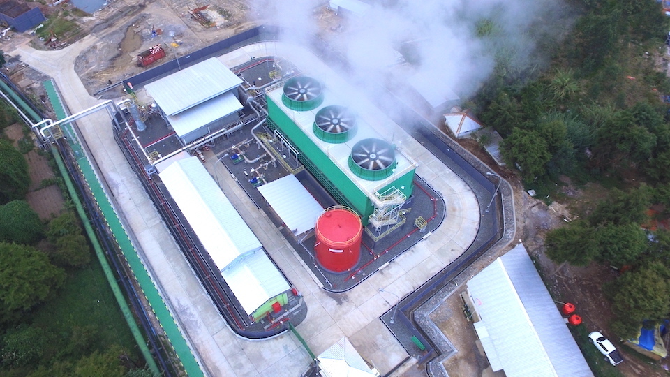 A view of the power plant, a large green and white building with three prominent circular structures on top where emissions of steam are coming from.