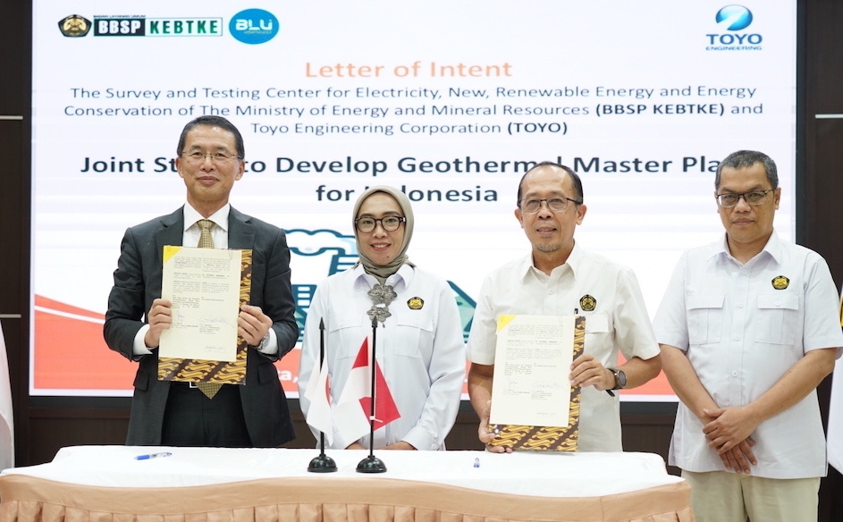 Toyo Engineering president and CEO, in a suit holding signed documents and woman wearing a hijab, and other two men in white shirts are standing behind a table with microphones.