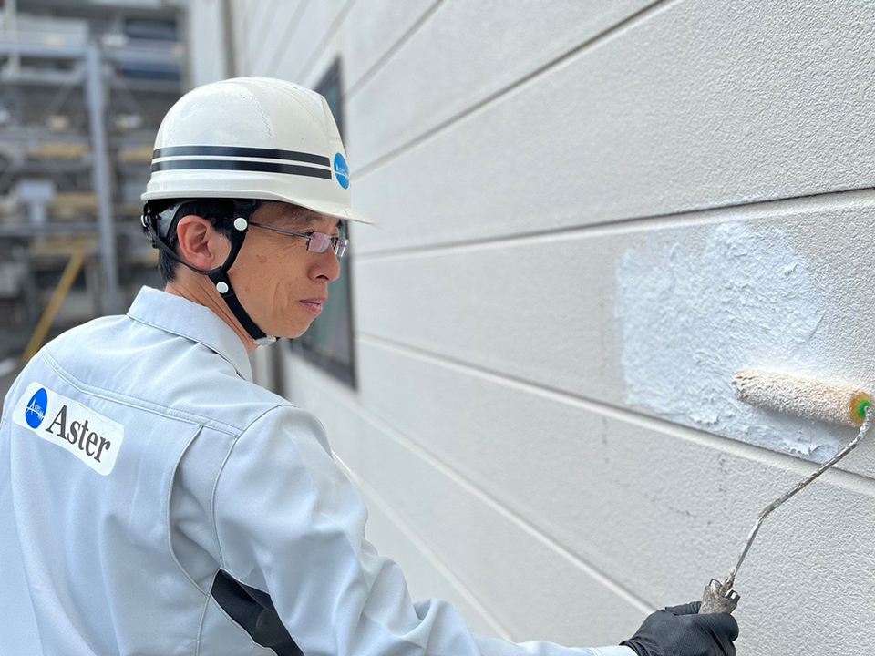 A person, wearing a white hard hat and a white uniform with a blue logo reading "Aster", painting an white wall with a paint roller.