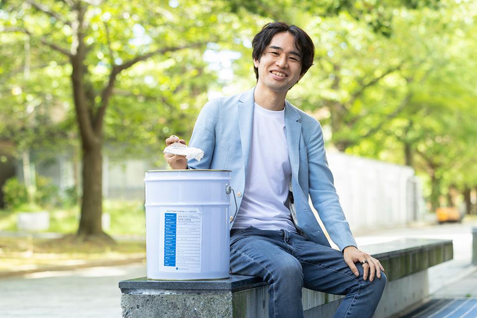 A person, smiling at the camera, sitting outdoors in what appears to be a park, next to him is a large white container with a blue label.