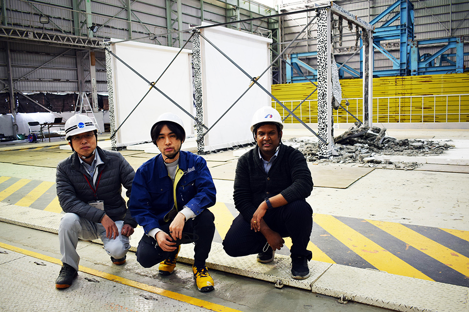 Three workers are crouched on a test site floor, two frames with white panels, and one with a broken panel in the background.