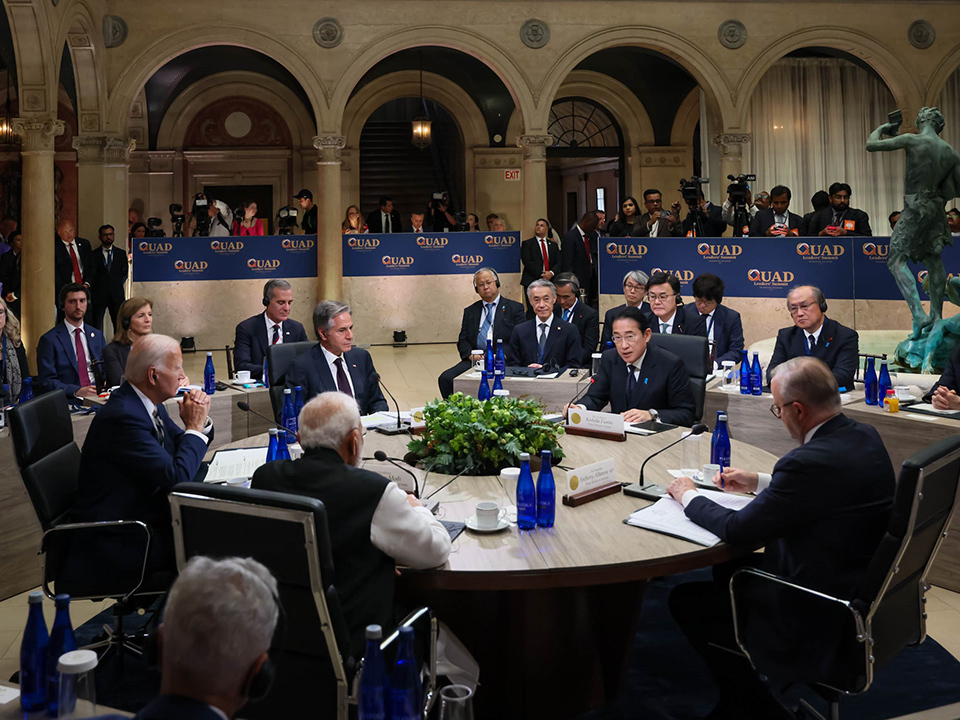 Officials are seated around a large circular table with blue water bottles and name placards visible.