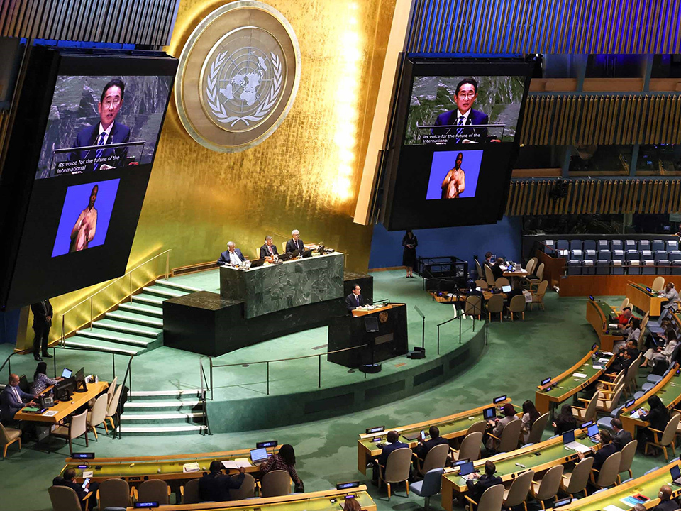 The United Nations General Assembly hall, showing its distinctive curved rows of delegate seating and the iconic UN emblem mounted on a golden wall.