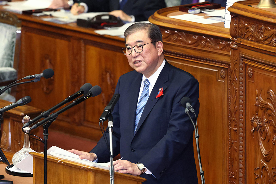 PM Ishiba,wearing a dark blue suit with glasses and a striped tie, stands at a wooden podium in the parliamentary chamber, addressing the assembly.