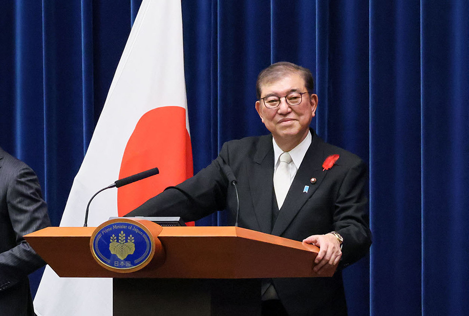 PM Ishiba,wearing a black suit with glasses and a white tie, stands and smiles in the press conference room.