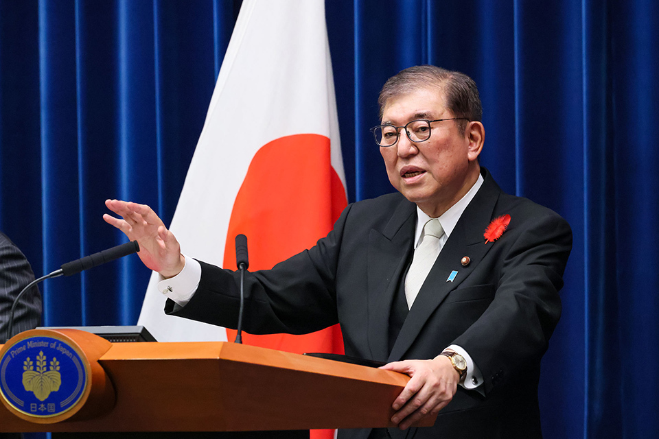 A man wearing a black suit and glasses is speaking at a podium with a Japanese flag in the background.