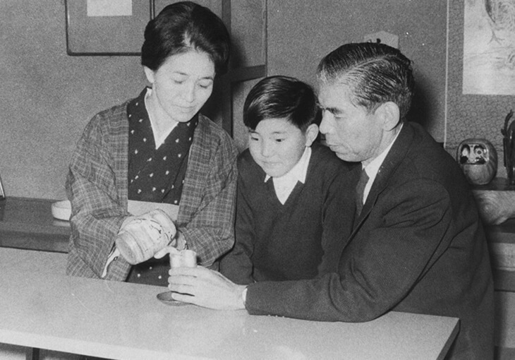 A woman pours a drink for a seated man and young boy at a table in a vintage setting.