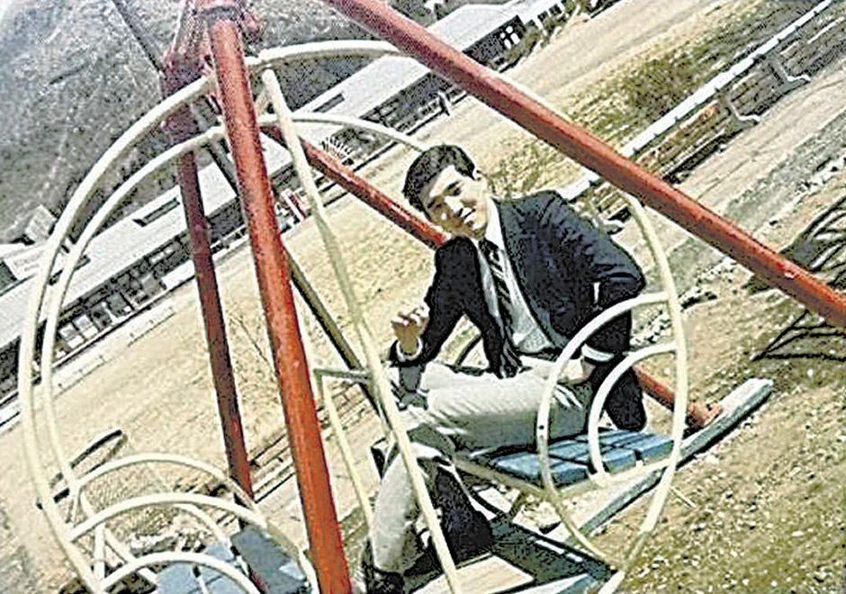 A man dressed in a formal suit is sitting on a playground swing that has a circular, metallic frame.