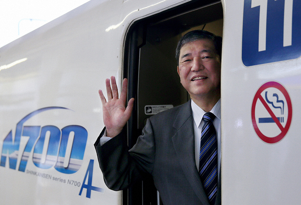 A man in a suit and tie waves cheerfully from the door of a train.