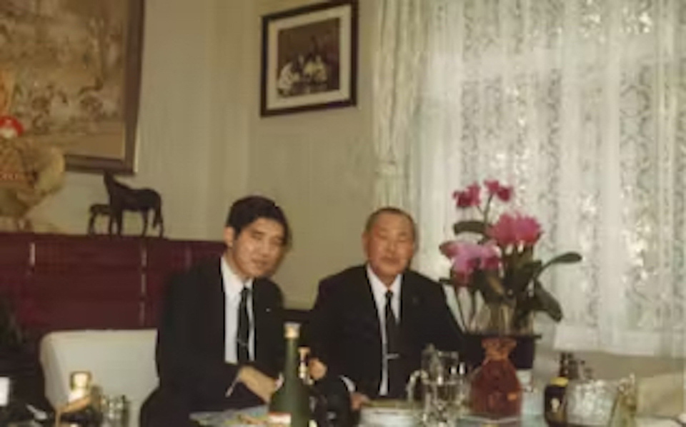 Two men in formal suits at a table set with various glasses, bottles, and a vase of pink flowers.