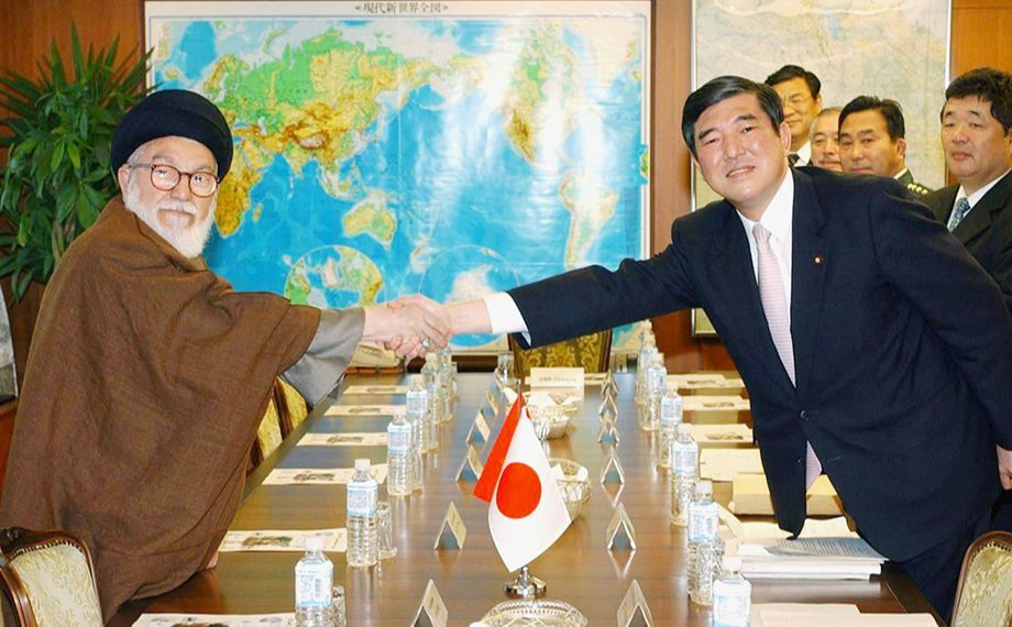 Two men shaking hands over a table, with a detailed wolrd map in the background.