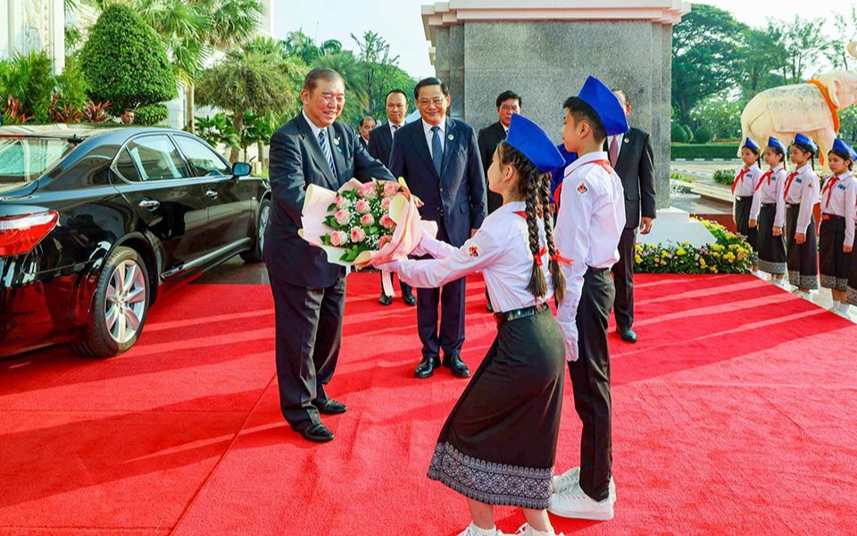 Prime Minister Ishiba receives a bouquet from two children in traditional uniforms on a red carpet, at a welcome ceremony in Lao PDR on October 11, 2024.