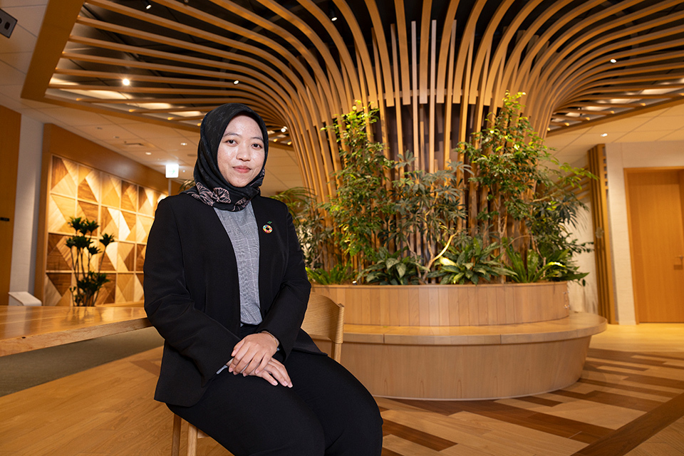 A woman wearing a black hijab, a black blazer over a grey top, and a ceiling with curved wooden slats create a radial pattern in the background.