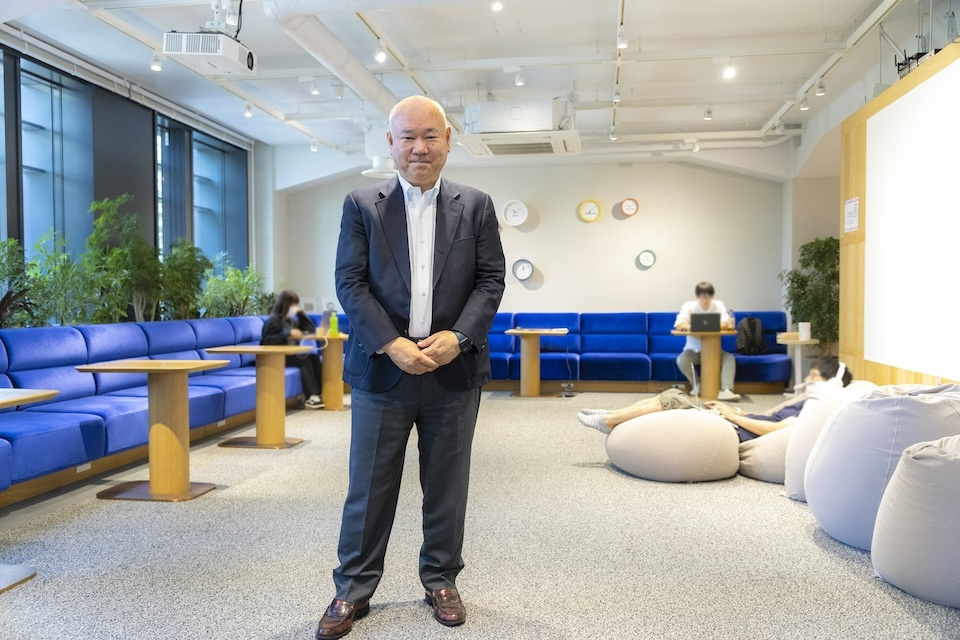 In an office setting, a professional person stands among blue chairs and white bean bags, highlighting a modern and corporate environment.