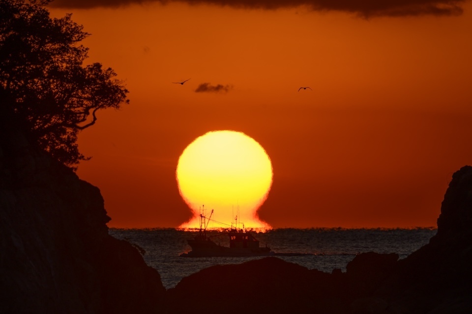 A large orange sun, resembling a Daruma, sets majestically over the calm ocean waters of Sukumo City.