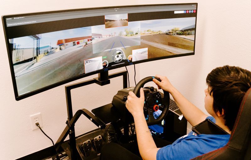 A man intently engages a large computer monitor in a well-lit room, remotely driving a rental car.