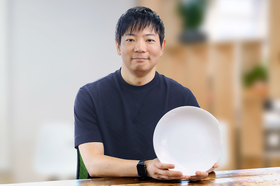 A man in a dark blue T-shirt sits at a table, holding a white plate in his hands.