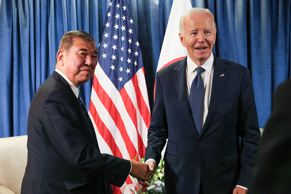 Two men shaking hands and two flags of Japan and the United States in the background.