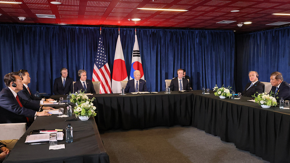 A diverse group of individuals seated at a table, with three flags displayed prominently in the background.