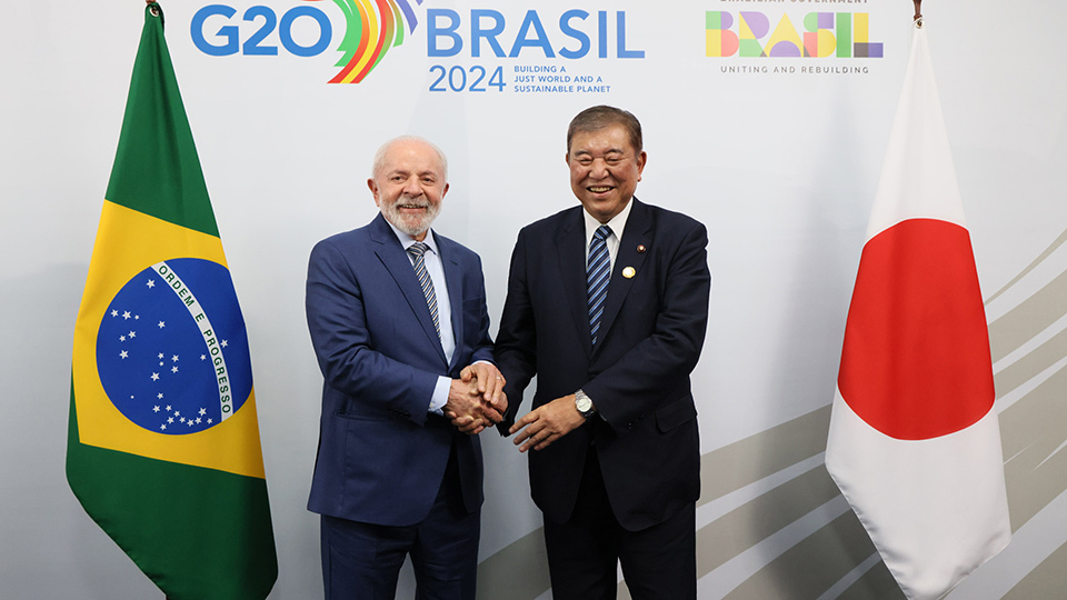 Two men engaged in a handshake, smiling, with Brazilian and Japanese flags displayed prominently in the background.