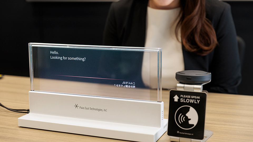 A clear display device showing 'Hello' text on a white base, with a 'Please Speak Slowly' microphone beside it. A smiling woman sits behind.