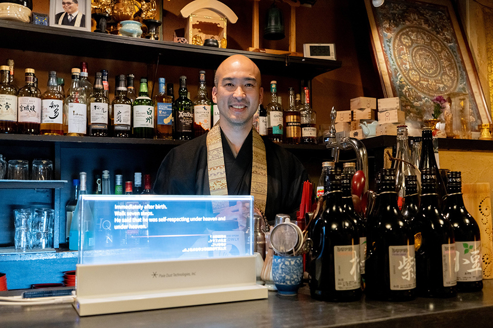 A smiling monk behind the VUEVO Display at VOWZ Bar, with Japanese whiskey bottles lining the shelves behind the counter.