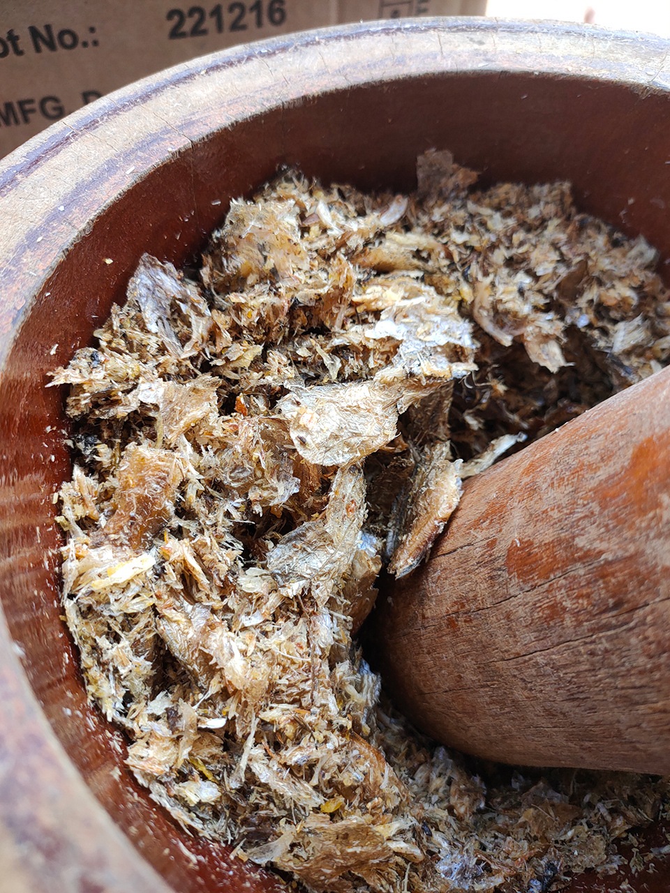 A wooden masher rests in a mortar containing finely ground dried fish, prepared for furikake seasoning.