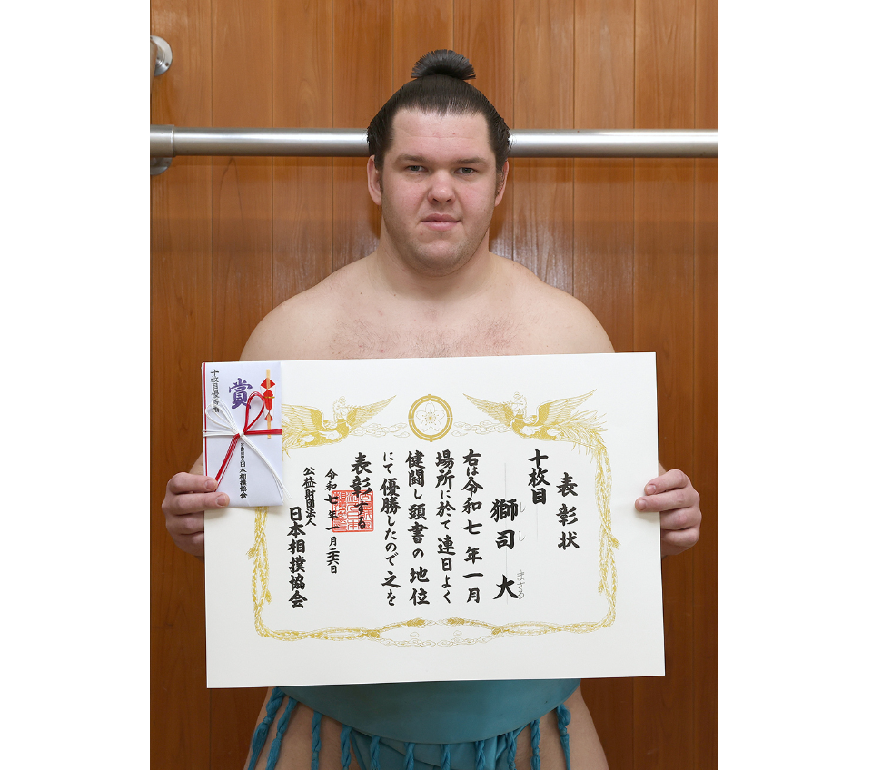 A man in sumo attire proudly holds a large Certificate of Merit.