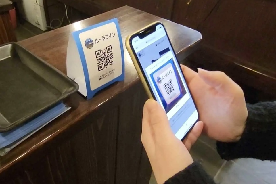 An individual holds a smartphone scanning a QR code of the "Rural Coin" at the cafe.